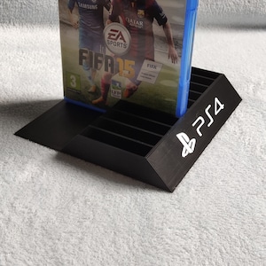 Peut inclure: Un support en plastique noir pour les boîtiers de jeux vidéo avec le logo PlayStation 4 sur le devant. Un boîtier de jeu PlayStation 4 bleu est dans le support. Le boîtier de jeu est pour FIFA 15 et présente Lionel Messi et Eden Hazard sur la couverture.