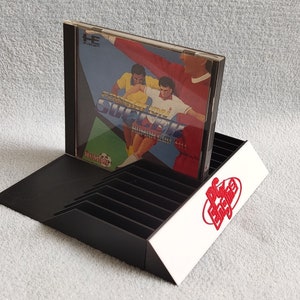 Peut inclure: Support de rangement noir en plastique pour jeux vidéo avec un panneau avant blanc. Le support comporte des fentes pour ranger les cartouches de jeux vidéo. Une cartouche de jeu vidéo est affichée dans la fente supérieure. La cartouche présente une image colorée de deux joueurs de football sur le devant. Le texte "Super Soccer" est imprimé sur le devant de la cartouche.