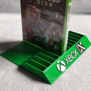 Peut inclure: Un support de jeu vidéo en plastique vert avec le logo Xbox Series X. Le support comporte des fentes pour plusieurs boîtiers de jeu. Un boîtier de jeu pour Watch Dogs Legion est affiché dans le support.