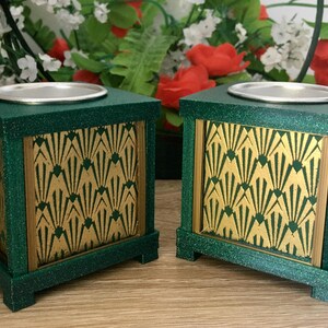 May include: Two green Art Deco candle holders with gold detailing. Each cube-shaped holder has a silver metal insert on top. The front panels feature a gold fan design. The candle holders are displayed on a wooden surface.