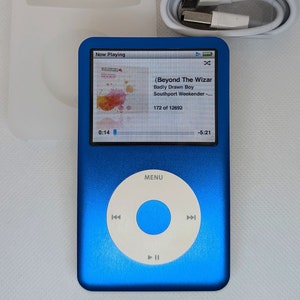 Puede incluir: Un iPod Apple azul con una pantalla blanca que muestra la canción "Beyond The Wizard" de Badly Drawn Boy. La pantalla también muestra que la canción es la 172 de 12692 y que la hora es 0:14 y -5:21. El iPod tiene un botón circular blanco con la palabra "MENU" en él.