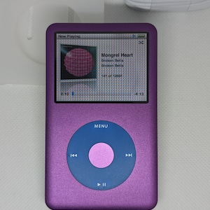 Puede incluir: Un iPod Nano morado con una pantalla blanca que muestra "Now Playing" y la canción "Mongrel Heart" de Broken Bells. La pantalla también muestra que la canción es la pista 141 de 12691. El iPod tiene un botón circular azul con un centro rosa y las palabras "MENU" en blanco.