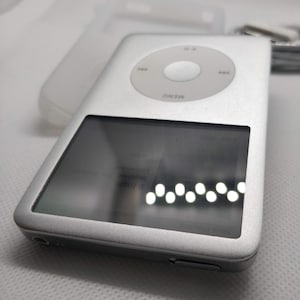 May include: A silver Apple iPod with a black and white screen displaying the text "Iron & Wine" and "The Trapeze Swinger". The iPod has a circular button with the word "MENU" printed on it.