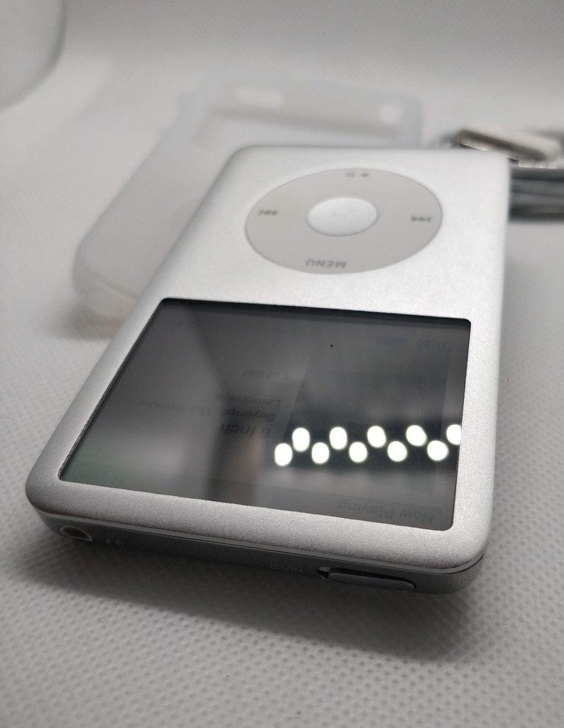 May include: A silver Apple iPod with a black and white screen displaying the text "Iron & Wine" and "The Trapeze Swinger". The iPod has a circular button with the word "MENU" printed on it.