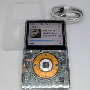 Puede incluir: Un reproductor de música digital transparente que revela componentes internos. La pantalla muestra "Now Playing" con información de la canción. También hay un cable de carga blanco y una funda protectora transparente.