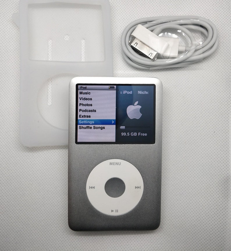 May include: A silver iPod with a black and white screen displaying the menu options: Music, Videos, Photos, Podcasts, Extras, Settings, and Shuffle Songs. The screen also shows "iPod Nicho" and "99.5 GB Free".