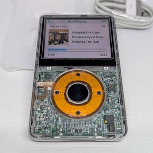 Puede incluir: Un reproductor MP3 transparente con una placa de circuito visible y un altavoz circular de color naranja. La pantalla muestra "Now Playing" con la canción "Bridging The Gap" de The Black Eyed Peas.