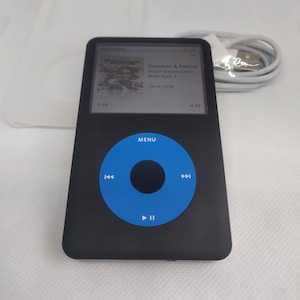 May include: A black iPod with a blue circular button in the center. The button has a play/pause symbol and arrows for navigating through music. The screen displays the song title "Common & Patrick" by Robert Glasper Experiment, "Black Radio 2", and the track number "195 of 12786".