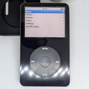 Puede incluir: Un iPod negro con una pantalla blanca que muestra las opciones de menú: Música, Fotos, Vídeos, Extras, Ajustes y Reproducción aleatoria. El iPod tiene un botón circular con la palabra "MENU" encima.
