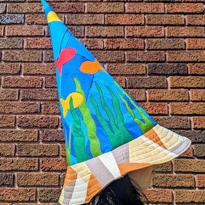 May include: A whimsical, handmade hat with a conical shape. The hat features a vibrant blue background with colorful fish and seaweed designs. The brim is a patchwork of various earth-toned fabrics, creating a unique and artistic accessory.