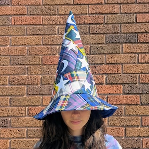 May include: A whimsical, wide-brimmed hat with a pointed crown. The hat features a patchwork design of plaid fabrics in blue, green, red, and yellow, accented with white stars. The brim is a solid blue, creating a unique, eye-catching accessory.
