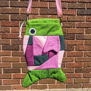 May include: A colorful fish-shaped crossbody bag with a pink strap. The bag features a patchwork design in shades of green, pink, and purple. The bag has a white drawstring closure and a large white eye. The bag is hanging in front of a brick wall.