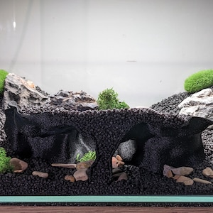 May include: An aquarium landscape featuring a black, cave-like structure made of porous material, surrounded by dark gravel. Green moss and small rocks are placed throughout the scene, creating a naturalistic underwater environment.
