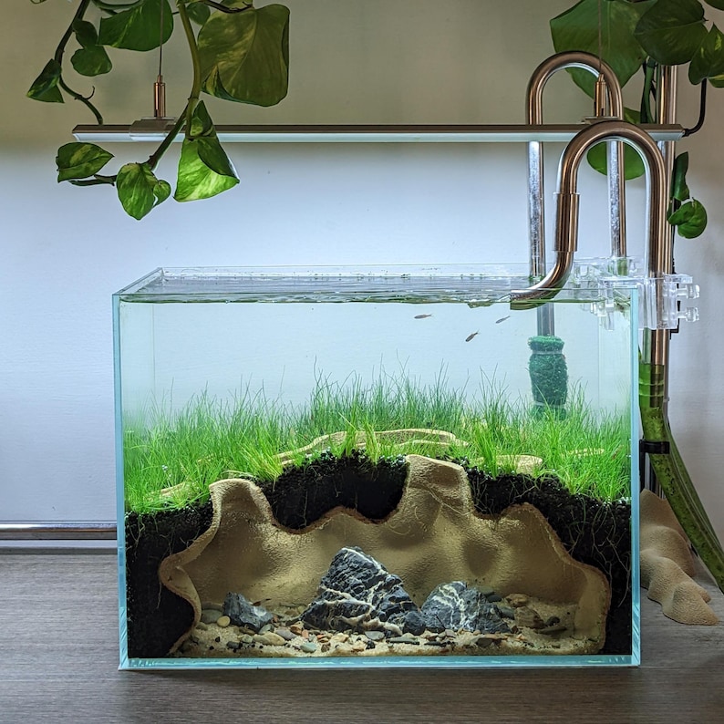 May include: A rectangular glass aquarium with a meticulously designed underwater landscape. The tank features vibrant green aquatic plants, beige rock formations, and dark gray stones. A metal light fixture arches over the top, and a trailing green plant is visible.