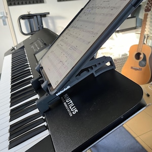 Può includere: Una tastiera nera con un supporto per tablet fissato sulla parte superiore. Il tablet mostra spartiti musicali. Il supporto è etichettato "Nautilus Music Workstation" e "Bmftudioent".