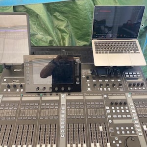 Könnte beinhalten: Ein schwarzes digitales Mischpult Behringer WING mit einem zentralen Touchscreen, mehreren Fadern und Drehreglern. Ein Tablet ist auf der linken Seite montiert und ein silberner Laptop befindet sich auf der rechten Seite des Mischpults.