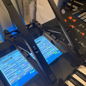 May include: A black keyboard with a blue screen displaying a list of songs. The screen shows song titles such as "24K Magic", "Africa", "Can't Feel My Face", "Jump", "Radio Gaga", and "One Vision".