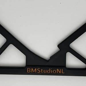 May include: Black plastic tool with a geometric design, featuring a central V-shape and angled supports. The text "BMStudioNL" is printed in a copper color along the bottom edge. The tool appears to be a jig or a guide.