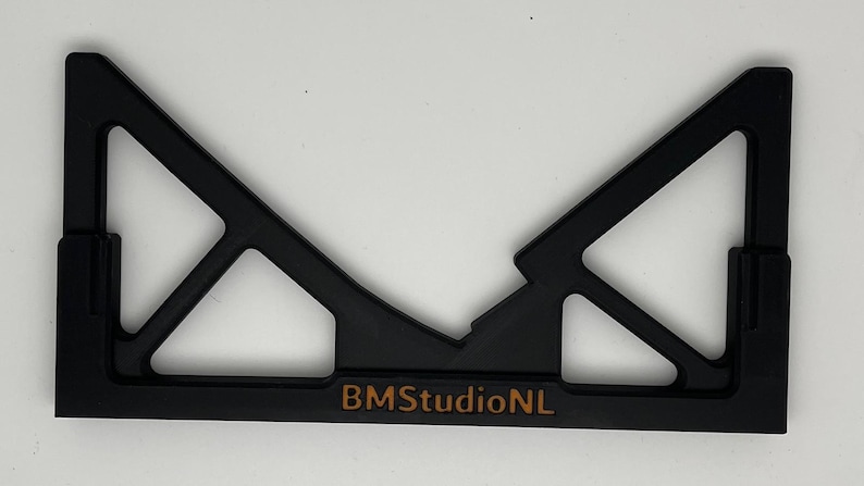 May include: Black plastic tool with a geometric design, featuring a central V-shape and angled supports. The text "BMStudioNL" is printed in a copper color along the bottom edge. The tool appears to be a jig or a guide.