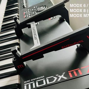 Puede incluir: Soporte de teclado negro y rojo con el texto "BMStudioNL" y "MODX M7". El soporte está diseñado para sostener un sintetizador de música. El teclado tiene teclas blancas y negras.