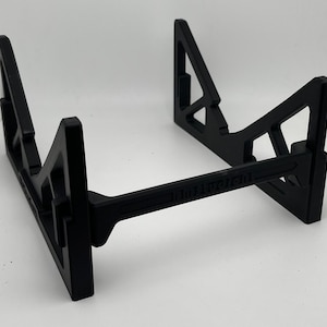 May include: Black, geometric stand with a horizontal support beam. The stand has a triangular design on the sides and a rectangular base. The text "101011101" is printed on the horizontal beam. The stand is designed to hold an item.