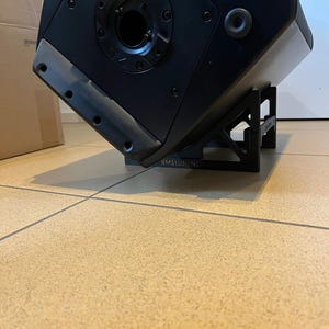 May include: A black speaker with a unique angular design, mounted on a black stand. The speaker has a circular opening and several screw holes. The text "V75" is visible on the speaker. The stand has the text "BMStudioNL".