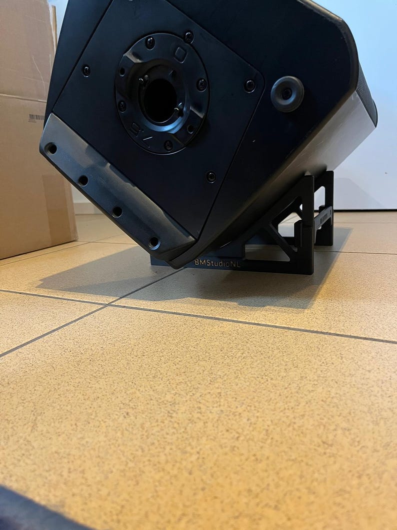 May include: A black speaker with a unique angular design, mounted on a black stand. The speaker has a circular opening and several screw holes. The text "V75" is visible on the speaker. The stand has the text "BMStudioNL".
