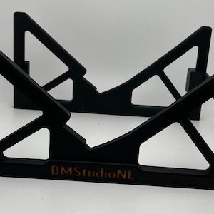 May include: Black wooden stand with a geometric design. The stand features a series of angled supports and a flat base. The text "BMStudioNL" is printed on the base. The stand is designed to hold an object.
