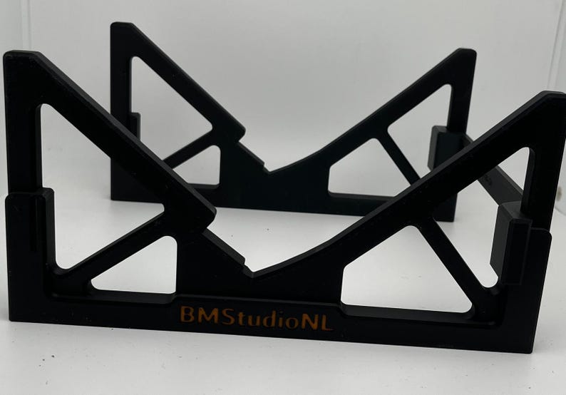 May include: Black wooden stand with a geometric design. The stand features a series of angled supports and a flat base. The text "BMStudioNL" is printed on the base. The stand is designed to hold an object.