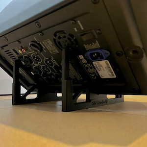 May include: A black audio speaker is supported by a black stand. The speaker's back panel features various input ports, power connections, and a label. The stand has a cross-braced design for stability and the text "3MStudioNL".