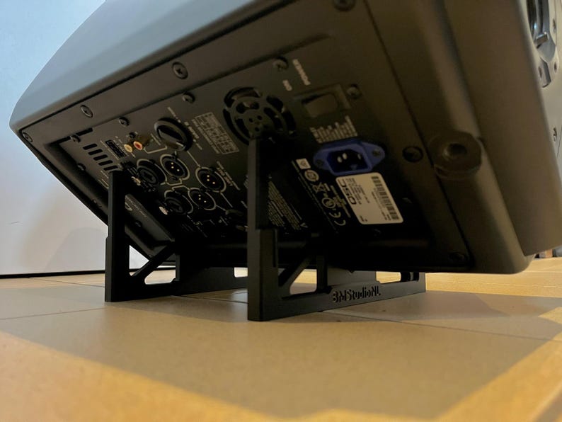 May include: A black audio speaker is supported by a black stand. The speaker's back panel features various input ports, power connections, and a label. The stand has a cross-braced design for stability and the text "3MStudioNL".