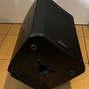 May include: A black, trapezoidal speaker with a textured, perforated front panel. The speaker has a glossy finish and a circular opening on the bottom. The brand name "QSC" is visible on the side.