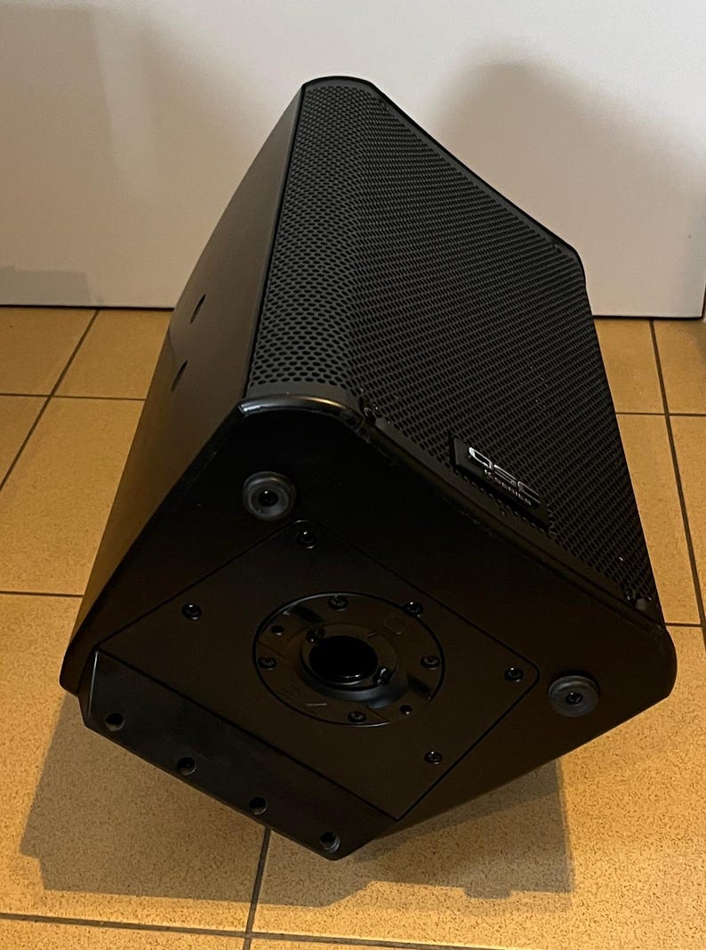 May include: A black, trapezoidal speaker with a textured, perforated front panel. The speaker has a glossy finish and a circular opening on the bottom. The brand name "QSC" is visible on the side.
