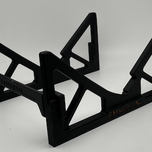 May include: A black, geometric stand with a triangular design. The stand has a rectangular base and angled supports, with the text "BMStudioNL" visible. The stand is designed to hold an item.