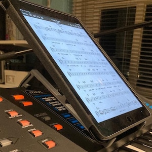 Può includere: Una tastiera nera con un tablet attaccato a un supporto. Il tablet mostra spartiti musicali. La tastiera ha una varietà di pulsanti e manopole, tra cui "Performance", "Utility", "Quick Setup", "Edit", "Compare", "Store", "Shift" e "Enter".