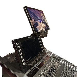 Peut inclure: Une console de mixage audio numérique Behringer noire avec un écran intégré et de nombreux boutons, molettes et curseurs. Un iPad est monté au-dessus de la console, affichant une image florale. La console est conçue pour le mixage audio professionnel.