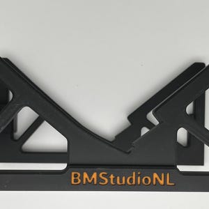 May include: Black, geometric shelf bracket with a triangular design. The bracket has a rectangular base with the text "BMStudioNL" in orange. The bracket is designed to hold items and is set against a white background.