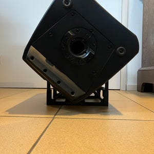 May include: A black speaker cabinet with a metal stand. The speaker has a circular opening in the center and is mounted on a black metal stand. The speaker is angled, revealing its side and bottom.