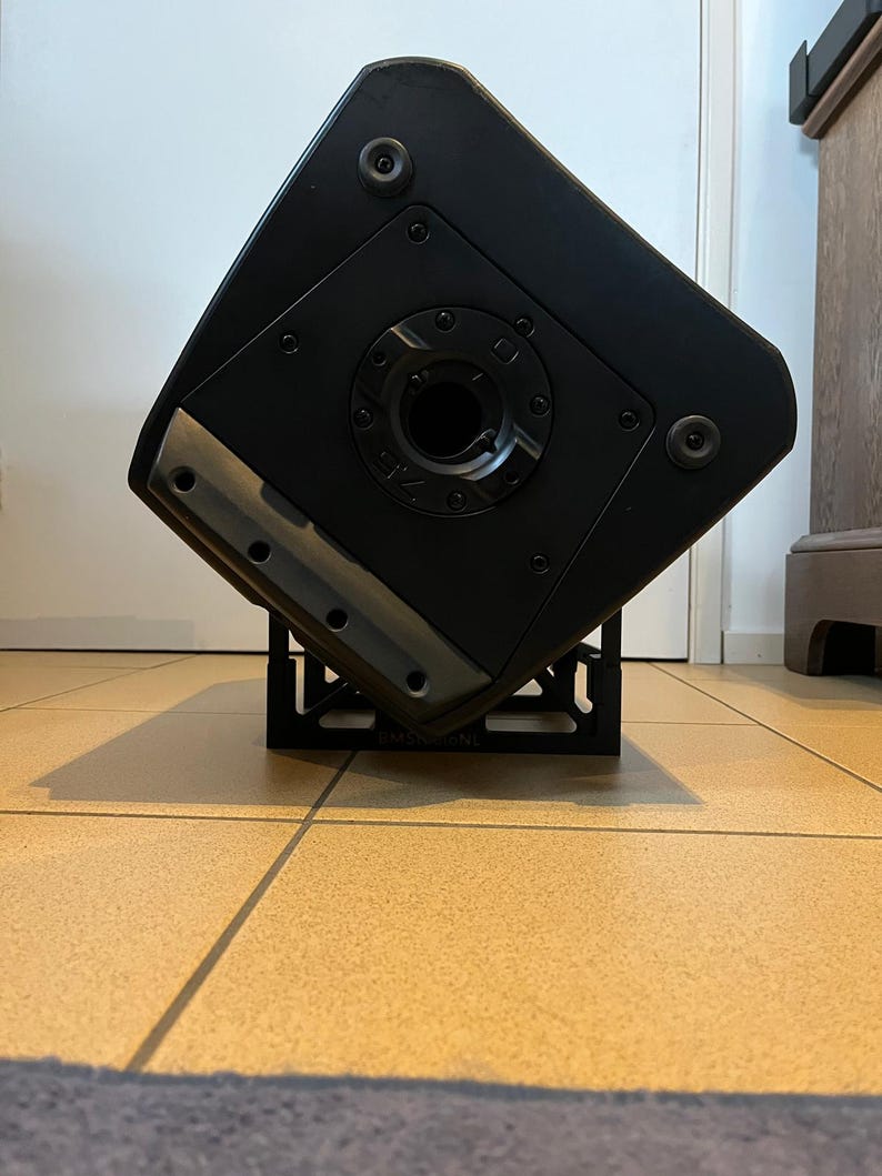 May include: A black speaker cabinet with a metal stand. The speaker has a circular opening in the center and is mounted on a black metal stand. The speaker is angled, revealing its side and bottom.