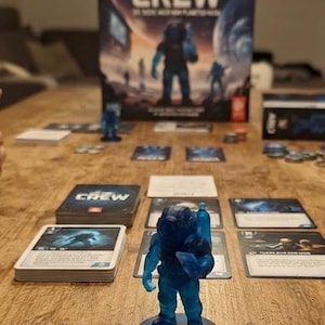 Könnte beinhalten: Ein Brettspiel mit dem Titel The Crew: Mission Deep Sea, das im Vordergrund eine blaue, durchscheinende Astronautenfigur zeigt. Das Spiel enthält eine große Schachtel, mehrere Stapel Spielkarten und kleine Spielsteine, die auf der Oberfläche verteilt sind und das Thema Weltraumforschung aufgreifen.