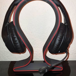 Könnte beinhalten: Ein schwarzes Headset mit einem roten Akzent am Kopfbügel liegt auf einem grauen Ständer mit einem roten Akzent. Der Ständer hat eine geschwungene Form und ein kleines Mikrofon ist am Headset befestigt.