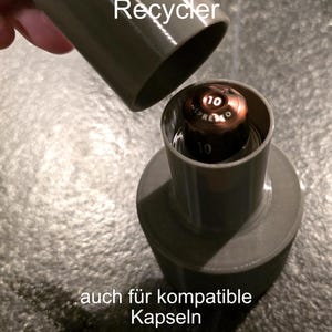 Könnte beinhalten: Ein grauer Nespresso Kapsel-Recycler mit dem Text "Nespresso Kapsel Recycler" und "auch für kompatible Kapseln". Der Recycler dient zum Halten und Sammeln gebrauchter Nespresso-Kapseln. Eine gebrauchte Kapsel ist sichtbar.