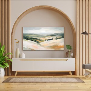 May include: A white media console with three drawers sits in front of a large framed painting of a landscape with rolling hills and a blue sky. The painting is hung in an arched doorway with wood trim.