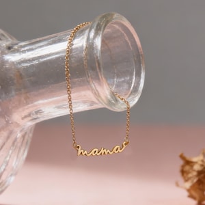 Könnte beinhalten: Eine zarte, goldfarbene Halskette mit dem Wort "mama" in Schreibschrift. Die Halskette ist über einer klaren Glasflasche drapiert, vor einem rosa-grauen Hintergrund. Die Kette ist fein und der Anhänger klein.