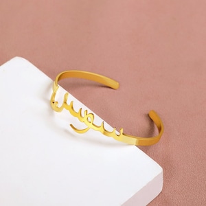 Könnte beinhalten: Goldfarbenes Armband mit dem arabischen Wort "سعيد" (bedeutet "glücklich") in einer stilisierten Schrift.