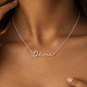 Könnte beinhalten: Eine silberfarbene Metallhalskette mit einer feinen Kette und einem zentralen Namensanhänger, der den Namen Olivia in Schreibschrift zeigt. Das Schmuckstück besteht aus einer dünnen, polierten Gliederkette und einem passenden Namensanhänger.