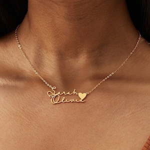 Op de afbeelding: Een goudkleurige ketting met de namen "Sarah Olivia" in cursief schrift en een kleine hartvormige bedel. De ketting hangt aan een fijne ketting. De achtergrond is een bruine top.