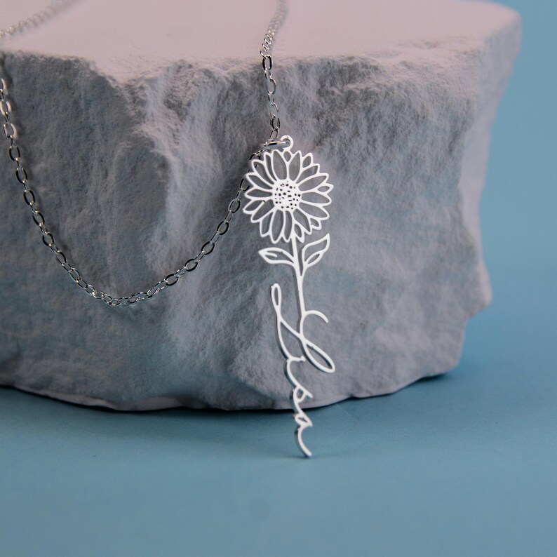 May include: A silver-colored necklace featuring a sunflower pendant with the name "Lisa" in a script font. The necklace is displayed on a gray, textured surface against a blue background. The sunflower pendant is intricately detailed.