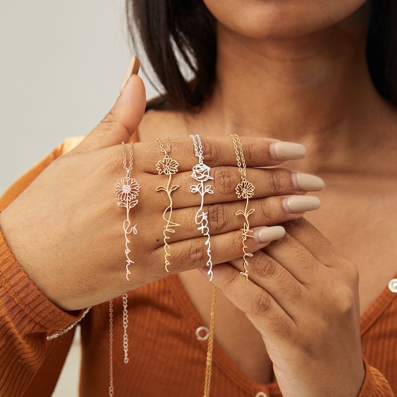 May include: Four floral pendant necklaces with personalized names. The necklaces feature a flower design at the top and a script name below. The necklaces are in silver and gold tones, with delicate chains.
