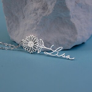 May include: A silver sunflower pendant necklace with the name "Lisa" in a cursive font. The necklace features a delicate sunflower design and is displayed against a blue background and a textured gray stone.